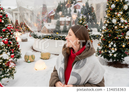 Smiling woman in warm winter jacket stands among decorated Christmas trees with red ornaments in a snowy Nutcracker-themed New Year installation Smiling woman in warm winter jacket stands among decorated Christmas trees with red ornaments in a snowy Nutcracker-themed New Year installation 134858363