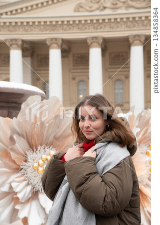 Woman stands near a snow-covered fountain with large white flower decorations in front of the Bolshoi Theatre facade and colonnade 134858364