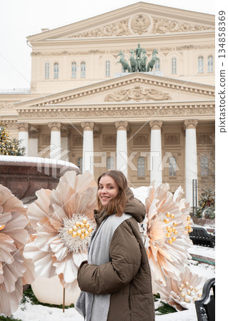 Woman stands near a snow-covered fountain with large white flower decorations in front of the Bolshoi Theatre facade and colonnade 134858369