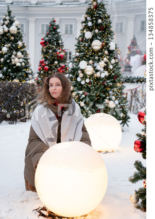 Woman in winter clothing touches glowing spherical light installation among decorated Christmas trees, snow, festive lights in modern holiday setting Woman in winter clothing touches glowing spherical light installation among decorated Christmas trees, snow, festive lights in modern holiday setting 134858375