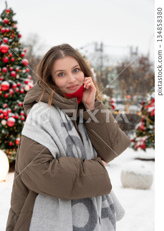 Young woman in warm winter jacket and scarf stands outdoors near decorated Christmas trees with red ornaments, snow, and soft festive lights Young woman in warm winter jacket and scarf stands outdoors near decorated Christmas trees with red ornaments, snow, and soft festive lights 134858380
