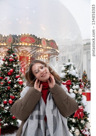 Smiling woman stands near decorated Christmas trees and a glowing carousel, surrounded by snow and warm festive lights in a cozy winter setting Smiling woman stands near decorated Christmas trees and a glowing carousel, surrounded by snow and warm festive lights in a cozy winter setting 134858383