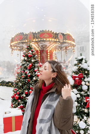 Woman in winter jacket and scarf stands near decorated Christmas trees and glowing carousel, touching her hair amid snow and warm festive lights Woman in winter jacket and scarf stands near decorated Christmas trees and glowing carousel, touching her hair amid snow and warm festive lights 134858388
