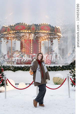 Smiling woman poses near glowing carousel, decorated Christmas trees, holding reusable mesh bag with mandarins in snowy festive setting 134858390