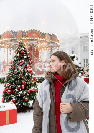 Woman in winter jacket and red sweater stands near decorated Christmas trees and glowing carousel, surrounded by snow and warm festive lights Woman in winter jacket and red sweater stands near decorated Christmas trees and glowing carousel, surrounded by snow and warm festive lights 134858391