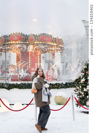 Smiling woman poses near glowing carousel, decorated Christmas trees, holding reusable mesh bag with mandarins in snowy festive setting Smiling woman poses near glowing carousel, decorated Christmas trees, holding reusable mesh bag with mandarins in snowy festive setting 134858402