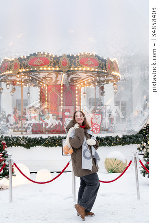 Smiling woman poses near glowing carousel, decorated Christmas trees, holding reusable mesh bag with mandarins in snowy festive setting Smiling woman poses near glowing carousel, decorated Christmas trees, holding reusable mesh bag with mandarins in snowy festive setting 134858403