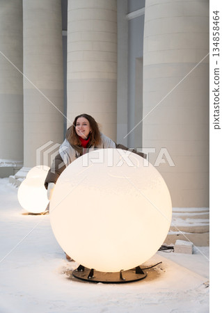 Smiling woman in winter clothing interacts with glowing spherical light installations in a snowy urban space, creating a modern festive winter mood 134858464