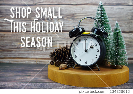 Festive holiday scene featuring a vintage alarm clock, pinecones, and miniature Christmas trees on a wooden base, with the text "Shop Small This Holiday Season" encouraging support for local businesse 134858613