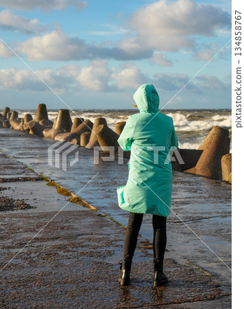 Woman in turquoise coat standing on stormy Baltic Sea pier with concrete wave breakers, rear view 134858767
