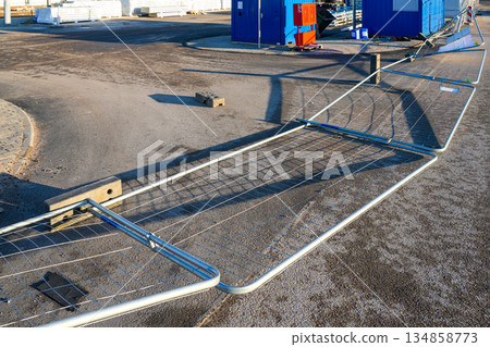 Wind-blown temporary construction site fence knocked down on asphalt near blue site containers 134858773