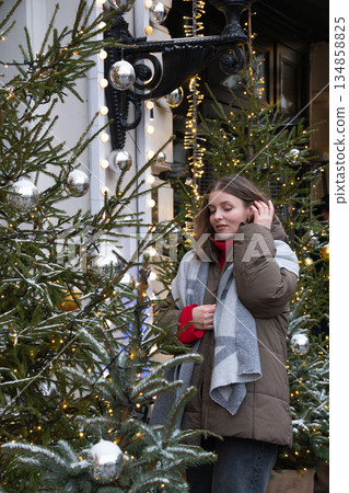 Woman in a winter coat and scarf stands among decorated fir trees with silver baubles and warm fairy lights, smiling in a cozy festive urban setting 134858825
