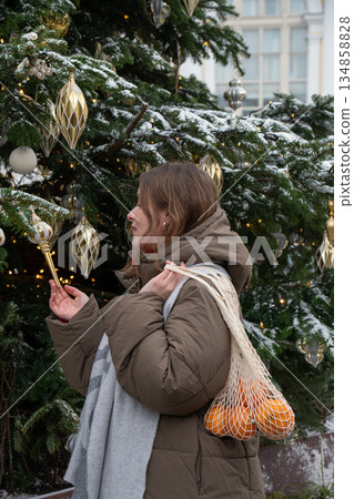 Woman in winter clothing holds reusable mesh bag with mandarins near decorated fir tree with snow, golden ornaments, warm festive lights 134858828