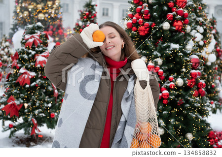 Joyful woman holds mandarin to her eye and laughs among decorated Christmas trees with snow, warm festive lights, creating playful holiday mood 134858832