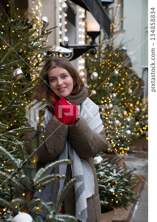 Woman in winter clothing smiles near a decorated Christmas tree with silver baubles, glowing fairy lights, soft golden bokeh in urban festive setting 134858834