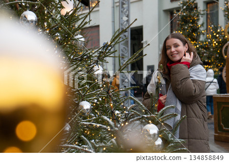 Woman in winter clothing smiles near a decorated Christmas tree with silver baubles, glowing fairy lights, soft golden bokeh in urban festive setting Woman in winter clothing smiles near a decorated Christmas tree with silver baubles, glowing fairy lights, soft golden bokeh in urban festive setting 134858849