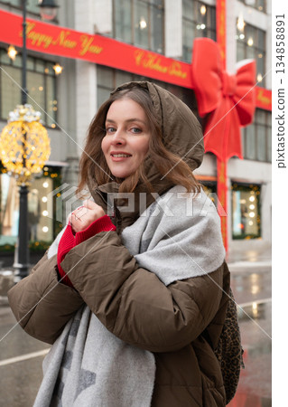 Woman in winter coat and scarf stands on wet street near decorated facade and entrance area of the TSUM Department Store during the New Year season 134858891