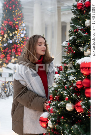 Woman in winter clothing stands close to decorated Christmas tree with red ornaments, snow, and warm festive lights, creating cozy holiday atmosphere Woman in winter clothing stands close to decorated Christmas tree with red ornaments, snow, and warm festive lights, creating cozy holiday atmosphere 134858897