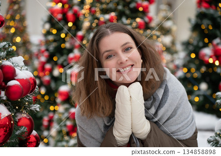 Woman in winter clothing rests her chin on gloved hands among decorated Christmas trees, snow, soft glowing lights, creating calm cozy holiday mood 134858898