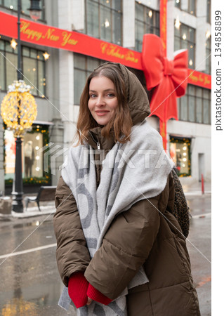 Woman in winter coat and scarf stands on wet street near decorated facade and entrance area of the TSUM Department Store during the New Year season 134858899