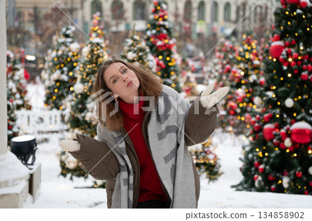 Playful woman in winter clothing shrugs and smiles among decorated Christmas trees with snow, glowing lights, creating cheerful, relaxed holiday mood 134858902