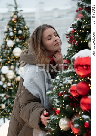 Woman in winter clothing stands close to decorated Christmas tree with red ornaments, snow, and warm festive lights, creating cozy holiday atmosphere 134858925
