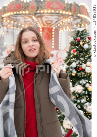 Woman in winter jacket adjusts her scarf near decorated Christmas trees and glowing carousel, surrounded by snow, warm festive lights 134858938