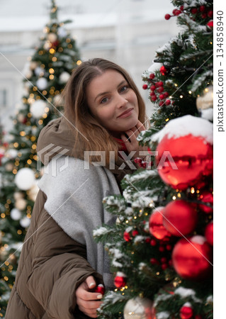 Woman in winter clothing stands close to decorated Christmas tree with red ornaments, snow, and warm festive lights, creating cozy holiday atmosphere Woman in winter clothing stands close to decorated Christmas tree with red ornaments, snow, and warm festive lights, creating cozy holiday atmosphere 134858940