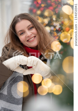 Smiling woman in winter clothing forms heart shape with her gloved hands, with soft golden light, decorated Christmas tree creating festive background 134858941