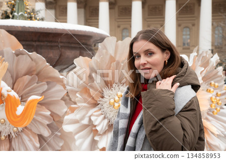 Woman in winter coat and scarf poses near large decorative flowers with soft beige tones, snow details, and calm festive mood Woman in winter coat and scarf poses near large decorative flowers with soft beige tones, snow details, and calm festive mood 134858953