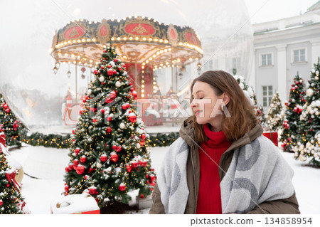 Woman in winter jacket and scarf stands near decorated Christmas trees and glowing carousel, touching her hair amid snow and warm festive lights 134858954