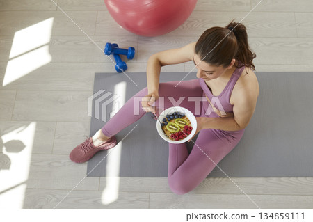 Young woman sitting on workout mat and eating healthy cereal with fruit and berries Young woman sitting on workout mat and eating healthy cereal with fruit and berries 134859111