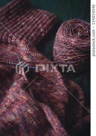 Unfinished melange brown sweater with knitting needles on dark green background. Knitting needles and balls of warm melange yarn. Knitting work in process. Knitting hobby. Knitwear concept. Unfinished melange brown sweater with knitting needles on dark green background. Knitting needles and balls of warm melange yarn. Knitting work in process. Knitting hobby. Knitwear concept. 134859346
