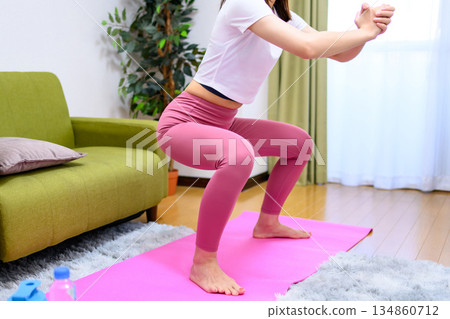 一位身穿瑜珈服的年輕女子在室內做深蹲，鍛鍊下半身，抬起臀部，進行塑形。 134860712