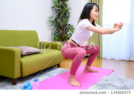 一位身穿瑜珈服的年輕女子在室內做深蹲,鍛鍊下半身,抬起臀部,進行塑形。 一位身穿瑜珈服的年輕女子在室內做深蹲,鍛鍊下半身,抬起臀部,進行塑形。 134860975