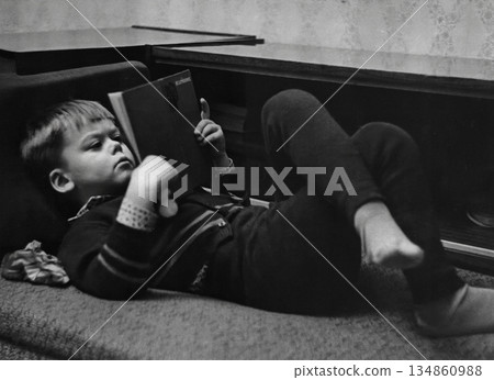 A vintage portrait of a six-year-old boy lying on a sofa reading a book. Retro photo from 1973. 134860988