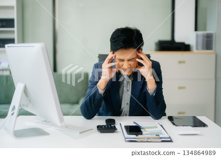 Frustrated young businessman working on a laptop computer sitting at his working place Frustrated young businessman working on a laptop computer sitting at his working place 134864989