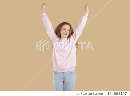 Curly redhead happy girl is rising her arms up on beige background and looking at camera. Curly redhead happy girl is rising her arms up on beige background and looking at camera. 134865137
