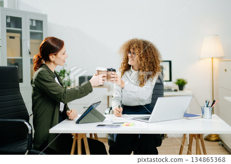 Two smiling women clinking coffee cups in a startup office, celebrating teamwork, friendship, and business success Two smiling women clinking coffee cups in a startup office, celebrating teamwork, friendship, and business success 134865368