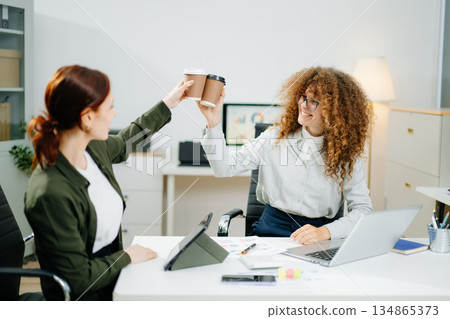 Two smiling women clinking coffee cups in a startup office, celebrating teamwork, friendship, and business success 134865373