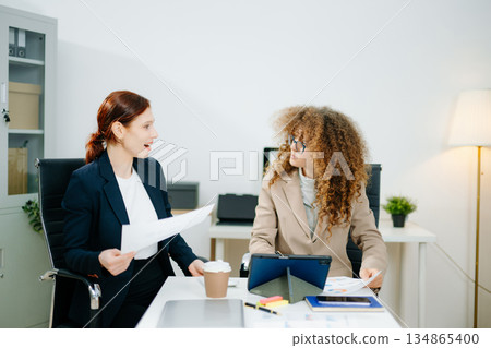 Two female professionals in smart casual outfits collaborate over finance charts and reports in a creative office space, planning business strategies 134865400