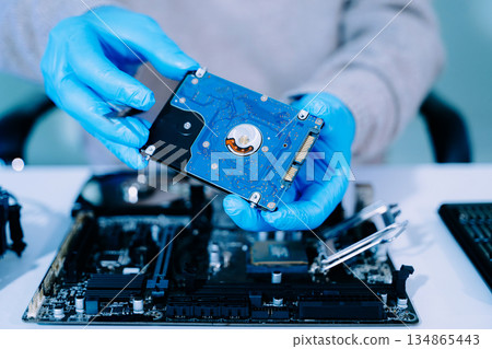 Closeup of technician with gloves holding blue hard drive circuit board above motherboard, symbolizing data recovery, storage, and computer repair. 134865443