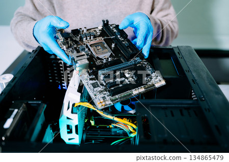 Closeup of technician with gloves holding motherboard above open PC case, symbolizing computer repair, upgrade, and technology innovation in IT service. Closeup of technician with gloves holding motherboard above open PC case, symbolizing computer repair, upgrade, and technology innovation in IT service. 134865479