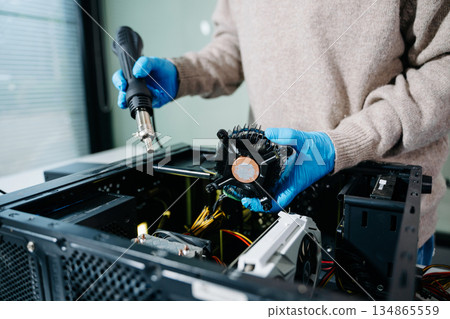 Closeup of technician in gloves holding CPU cooler and heat gun over desktop PC. Ideal image for computer repair, cooling system, and hardware Closeup of technician in gloves holding CPU cooler and heat gun over desktop PC. Ideal image for computer repair, cooling system, and hardware 134865559