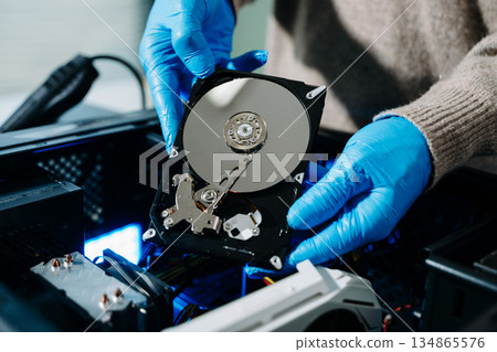 Closeup of gloved technician holding exposed hard drive above desktop PC. Ideal for data recovery, hardware repair, backup, and sensitive storage concepts 134865576