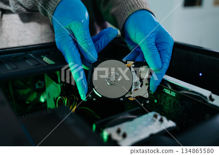 Closeup of gloved technician holding exposed hard drive above desktop PC. Ideal for data recovery, hardware repair, backup, and sensitive storage concepts Closeup of gloved technician holding exposed hard drive above desktop PC. Ideal for data recovery, hardware repair, backup, and sensitive storage concepts 134865580