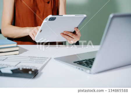 Creative female filmmaker working with tablet, clapperboard, and laptop in modern studio office. Film production, planning, and content creation scene. 134865588