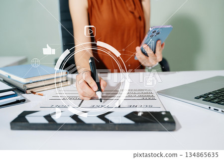 Creative woman working on tablet with digital icons and clapperboard. Modern media, marketing, and film production scene symbolizing innovation and content strategy. Creative woman working on tablet with digital icons and clapperboard. Modern media, marketing, and film production scene symbolizing innovation and content strategy. 134865635