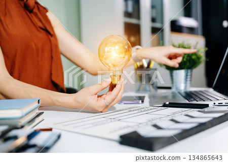 Creative woman holds a glowing lightbulb while using a laptop and clapperboard in modern office. Symbol of inspiration, film production, and innovative ideas. Creative woman holds a glowing lightbulb while using a laptop and clapperboard in modern office. Symbol of inspiration, film production, and innovative ideas. 134865643