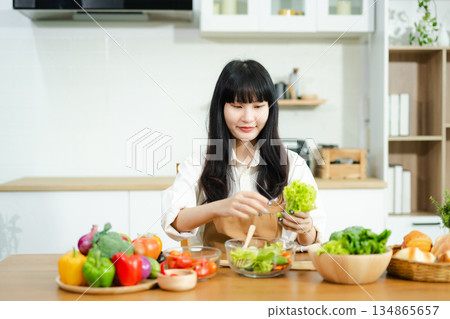Asian woman prepares healthy salad with fresh vegetables in a modern kitchen. Clean, vibrant, and perfect for nutrition, lifestyle, or home cooking concepts. Asian woman prepares healthy salad with fresh vegetables in a modern kitchen. Clean, vibrant, and perfect for nutrition, lifestyle, or home cooking concepts. 134865657